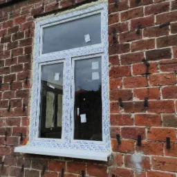 Nowe, białe okno z szybami zespolonymi, zamontowane w ceglanej ścianie budynku w Lublinie, zabezpieczone folią ochronną na ramie.