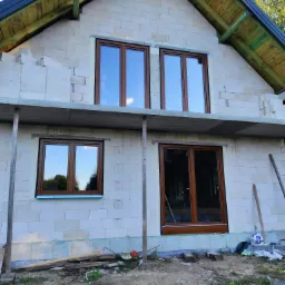 Surowy stan budynku w trakcie montażu stolarki okiennej z brązowymi ramami, widoczne rusztowania i materiały budowlane na zewnątrz.