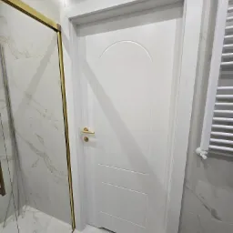 Białe drzwi wewnętrzne z zaokrąglonym panelem g&oacute;rnym i złotą klamką, umieszczone w łazience z marmurowymi płytkami i grzejnikiem drabinkowym.