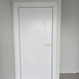 Welldoor Piotr Czerwonka - Białe, dwupanelowe drzwi wewnętrzne z zaokrąglonym górnym panelem i złotą klamką, zamontowane w szarej ścianie, widoczny fragment podłogi z szarych płytek imitujących marmur.