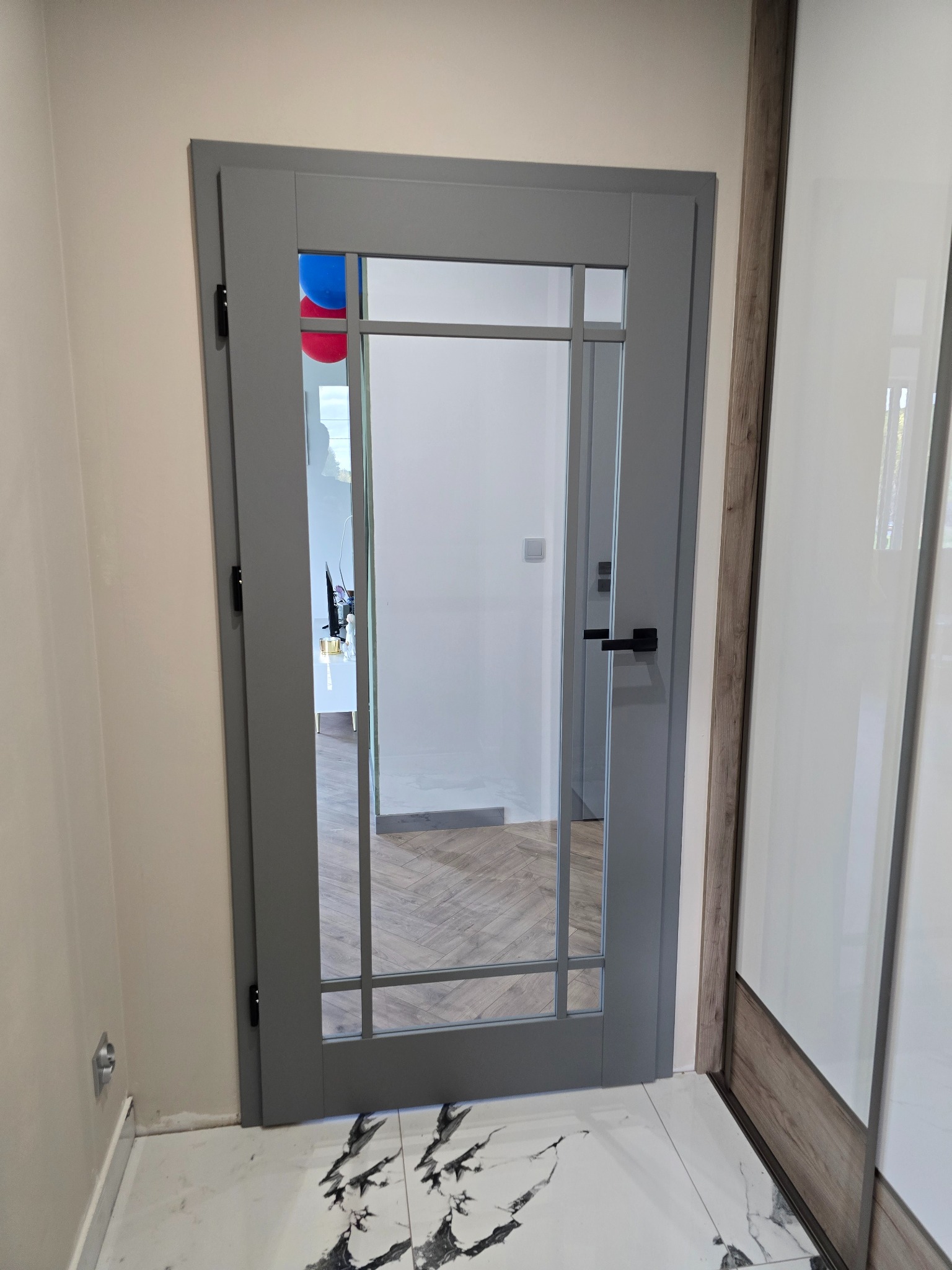 Szare drzwi wewnętrzne z szybą z podziałami, widok na sąsiednie pomieszczenie z parkietem w jodełkę i dekoracyjnymi balonami, na podłodze płytki imitujące marmur.