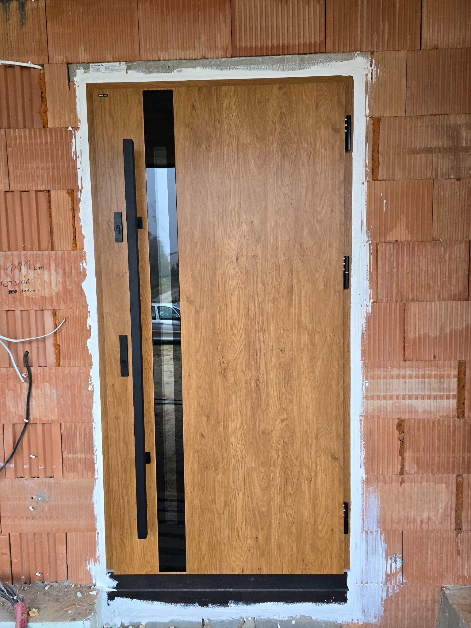 Montaż drzwi zewnętrznych w Lublinie: drzwi z drewnopodobną okleiną i długim, czarnym uchwytem, osadzone w surowym murze z cegły ceramicznej, z widocznymi śladami zaprawy.