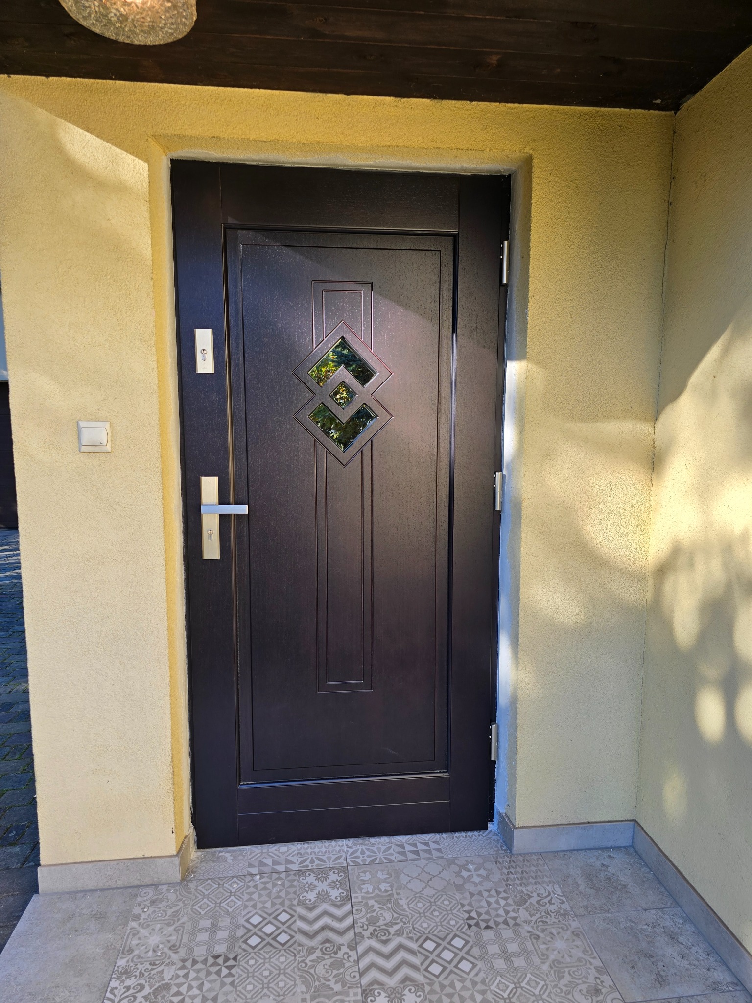 Ciemnobrązowe drzwi wejściowe z romboidalnym przeszkleniem, srebrna klamka i zamek, osadzone w żółtej elewacji, widoczne z zewnątrz na tle wzorzystych płytek podłogowych.