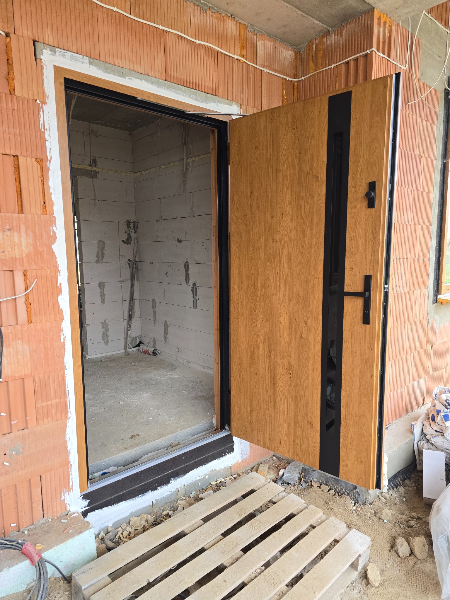 Montaż zewnętrznych drzwi wejściowych z drewnopodobną okleiną i czarnymi elementami wykończeniowymi w surowym stanie budynku z widocznymi bloczkami ceramicznymi i paletą na zewnątrz.