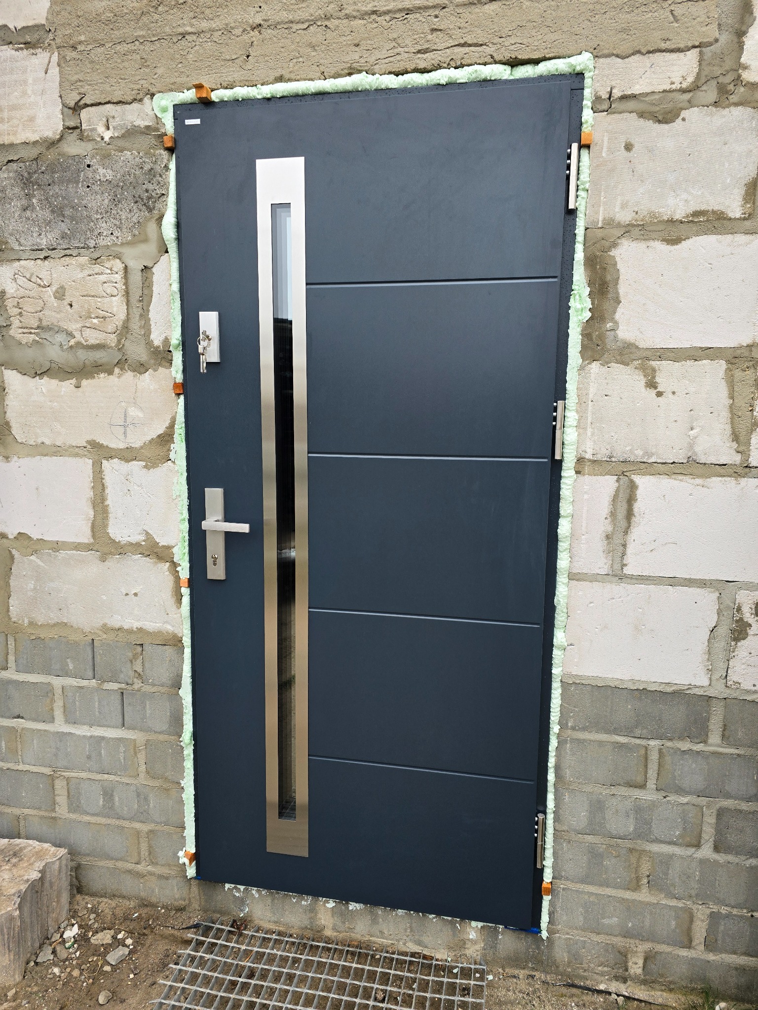 Montaż nowoczesnych, grafitowych drzwi zewnętrznych z przeszkleniem, widoczne uszczelnienie pianką montażową w surowym murze z betonu komórkowego.