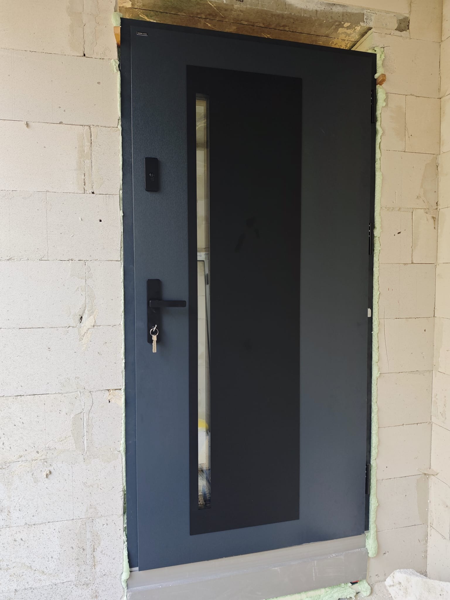 Szare drzwi zewnętrzne z szybą, zamontowane w surowej ścianie z betonu komórkowego, uszczelnione pianką montażową, widoczny zamek i klucze.