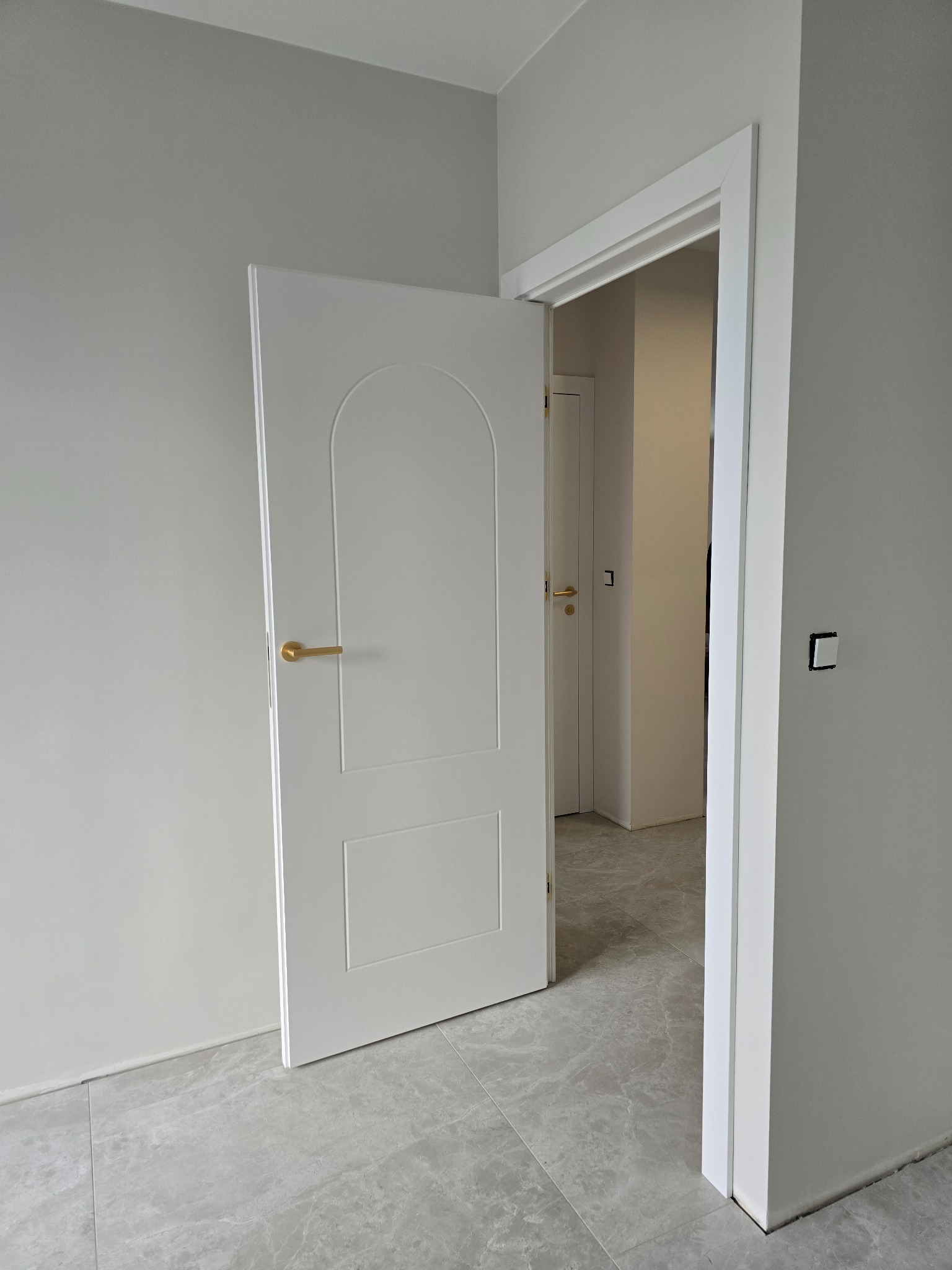 Otwarta, biała, wewnętrzna drzwi z zaokrąglonym panelem górnym i złotą klamką, widok na sąsiednie pomieszczenie z kolejnymi drzwiami, szare ściany i podłoga z płytek imitujących marmur.