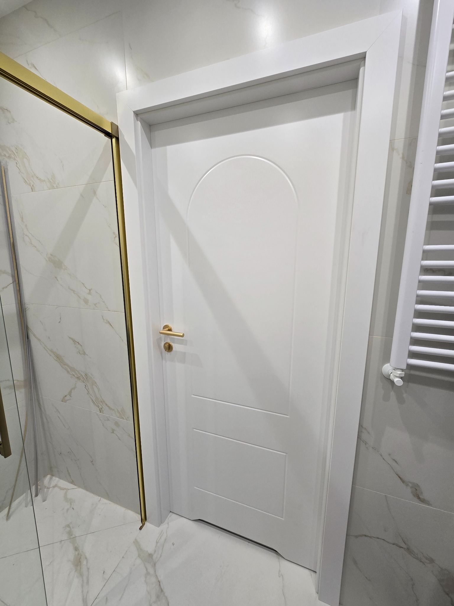 Białe drzwi wewnętrzne z zaokrąglonym panelem górnym i złotą klamką, umieszczone w łazience z marmurowymi płytkami i grzejnikiem drabinkowym.