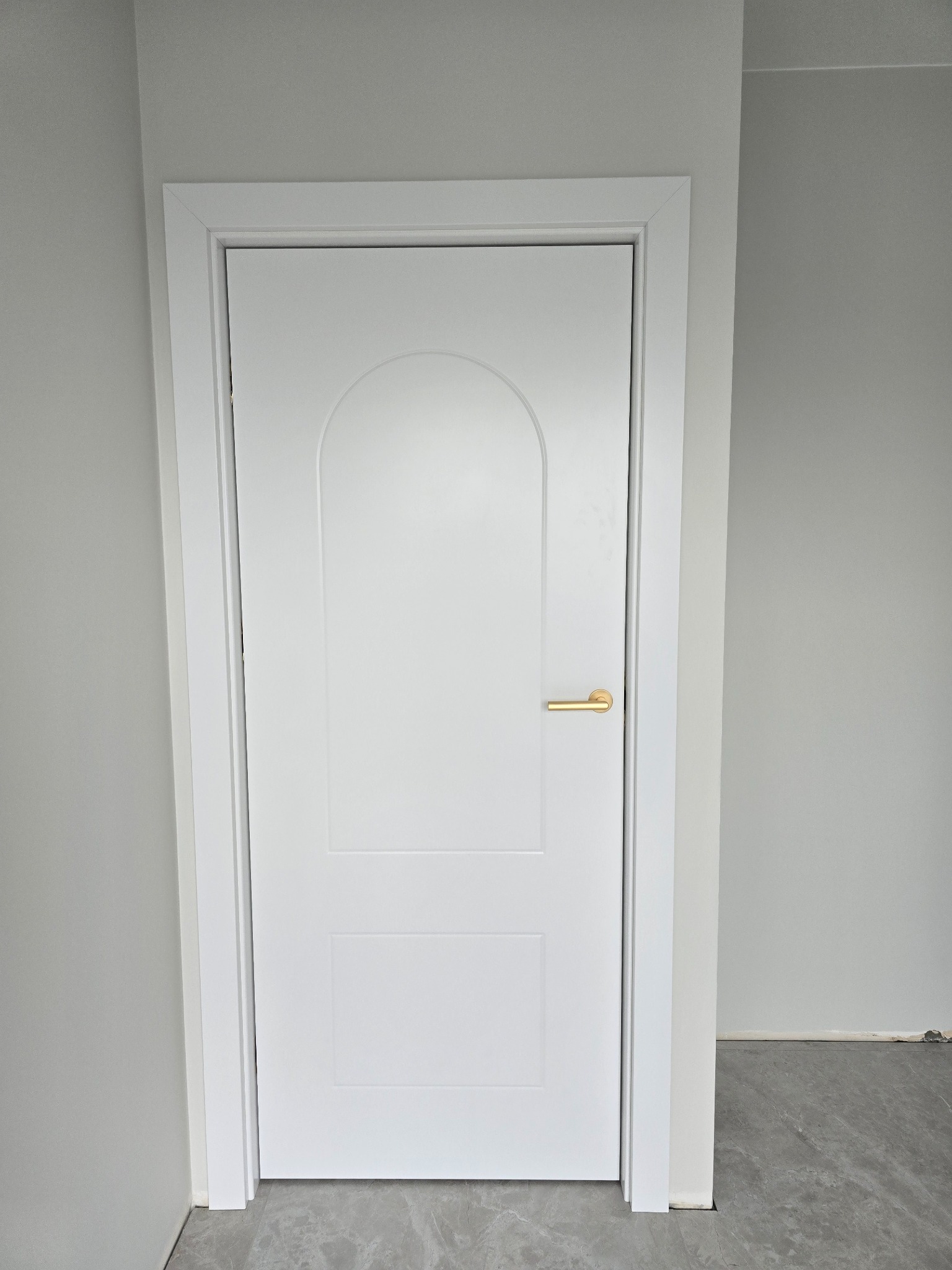 Białe, dwupanelowe drzwi wewnętrzne z zaokrąglonym górnym panelem i złotą klamką, zamontowane w szarej ścianie, widoczny fragment podłogi z szarych płytek imitujących marmur.