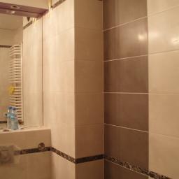 Mirbud - Fragment wykończonej łazienki z beżowo-szarymi płytkami na ścianach, białym grzejnikiem drabinkowym z żółtą gąbką i lustrem, w którym widać osobę i butelki wody mineralnej.