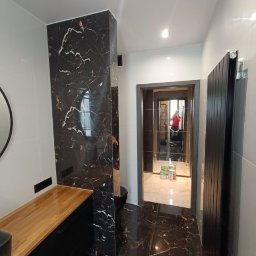 Mirbud - Wykończona łazienka z czarnymi płytkami imitującymi marmur na ścianach i podłodze, drewniany blat umywalki, czarny grzejnik i widoczne odbicie osoby w lustrze.