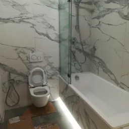 Nowoczesna łazienka z marmurowymi płytkami, białą wanną z parawanem, podświetleniem LED i podwieszaną toaletą. Na podłodze kartony zabezpieczające.