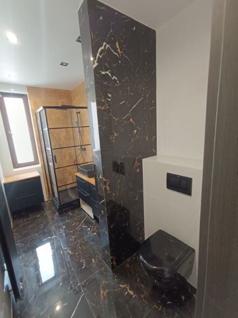 Wykończona łazienka z czarną armaturą i płytkami imitującymi marmur, kabina prysznicowa ze złotymi detalami, wisząca toaleta z czarnym przyciskiem spłukiwania.