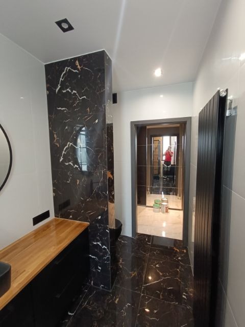 Wykończona łazienka z czarnymi płytkami imitującymi marmur na ścianach i podłodze, drewniany blat umywalki, czarny grzejnik i widoczne odbicie osoby w lustrze.