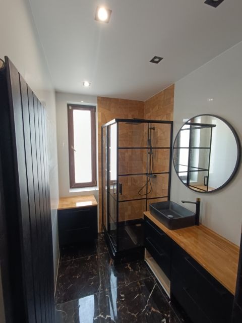 Nowoczesna łazienka z czarnymi meblami, kabiną prysznicową typu walk-in z czarnymi ramami, okrągłym lustrem i marmurową podłogą w odcieniach brązu i czerni.