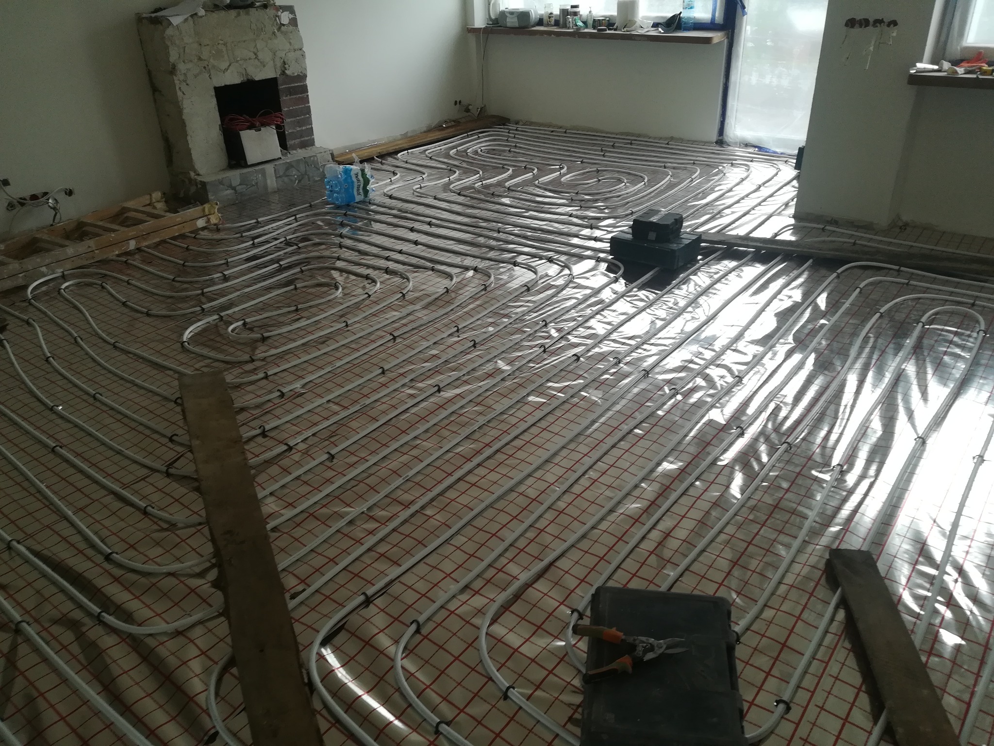 Rozłożone białe rury ogrzewania podłogowego na folii z czerwoną siatką w trakcie prac, widoczne narzędzia i elementy wykończenia wnętrza, takie jak kominek w surowym stanie i okna z zasłoniętą...