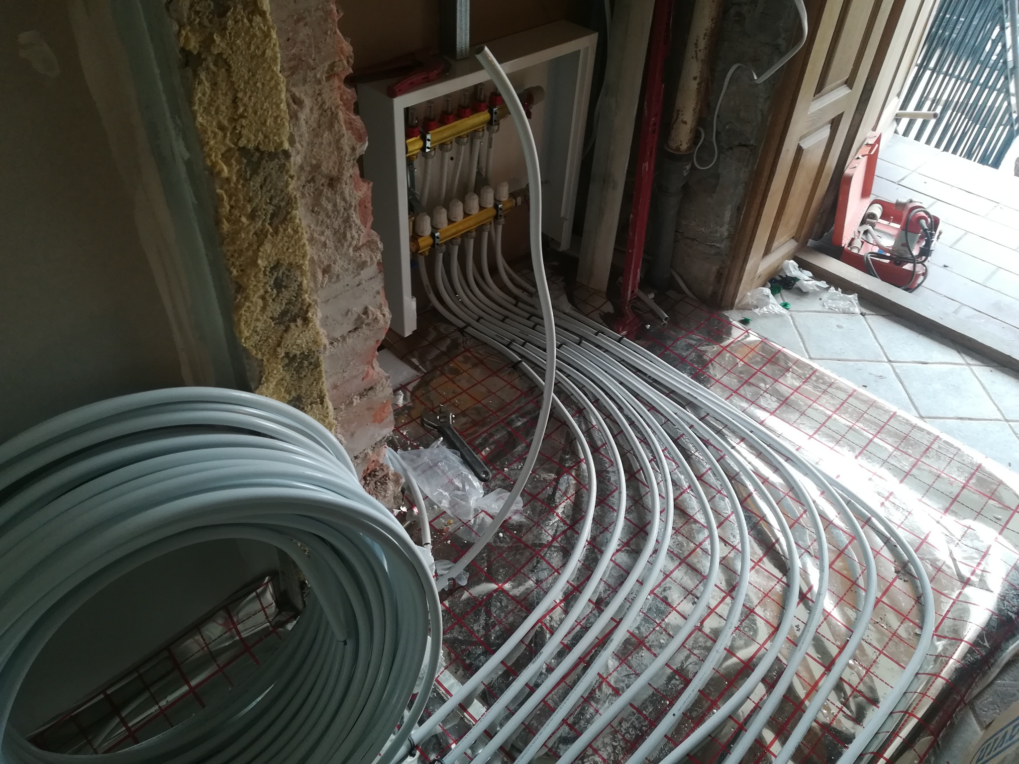 Instalacja ogrzewania podłogowego z rozdzielaczem na ścianie, widoczne rury w izolacji termicznej oraz narzędzia na podłodze w trakcie montażu.