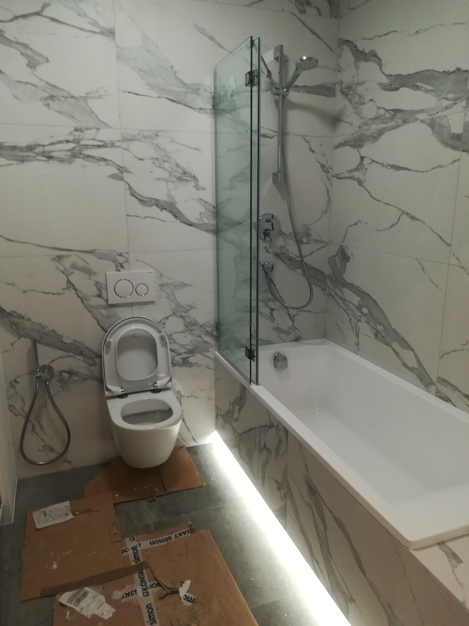 Nowoczesna łazienka z marmurowymi płytkami, białą wanną z parawanem, podświetleniem LED i podwieszaną toaletą. Na podłodze kartony zabezpieczające.