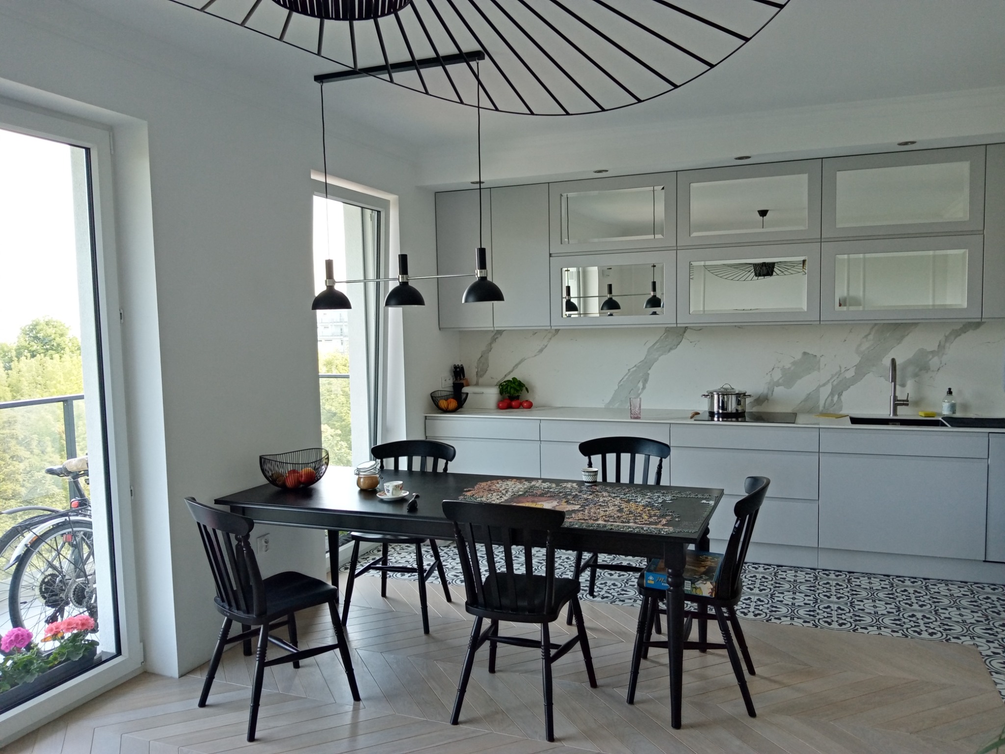 Jasne wnętrze kuchni z jadalnią: szare szafki kuchenne z lustrzanymi frontami, czarny stół z układanką, czarne krzesła, wzorzysta podłoga i drewniana podłoga w jodełkę, nowoczesna lampa wisząca...