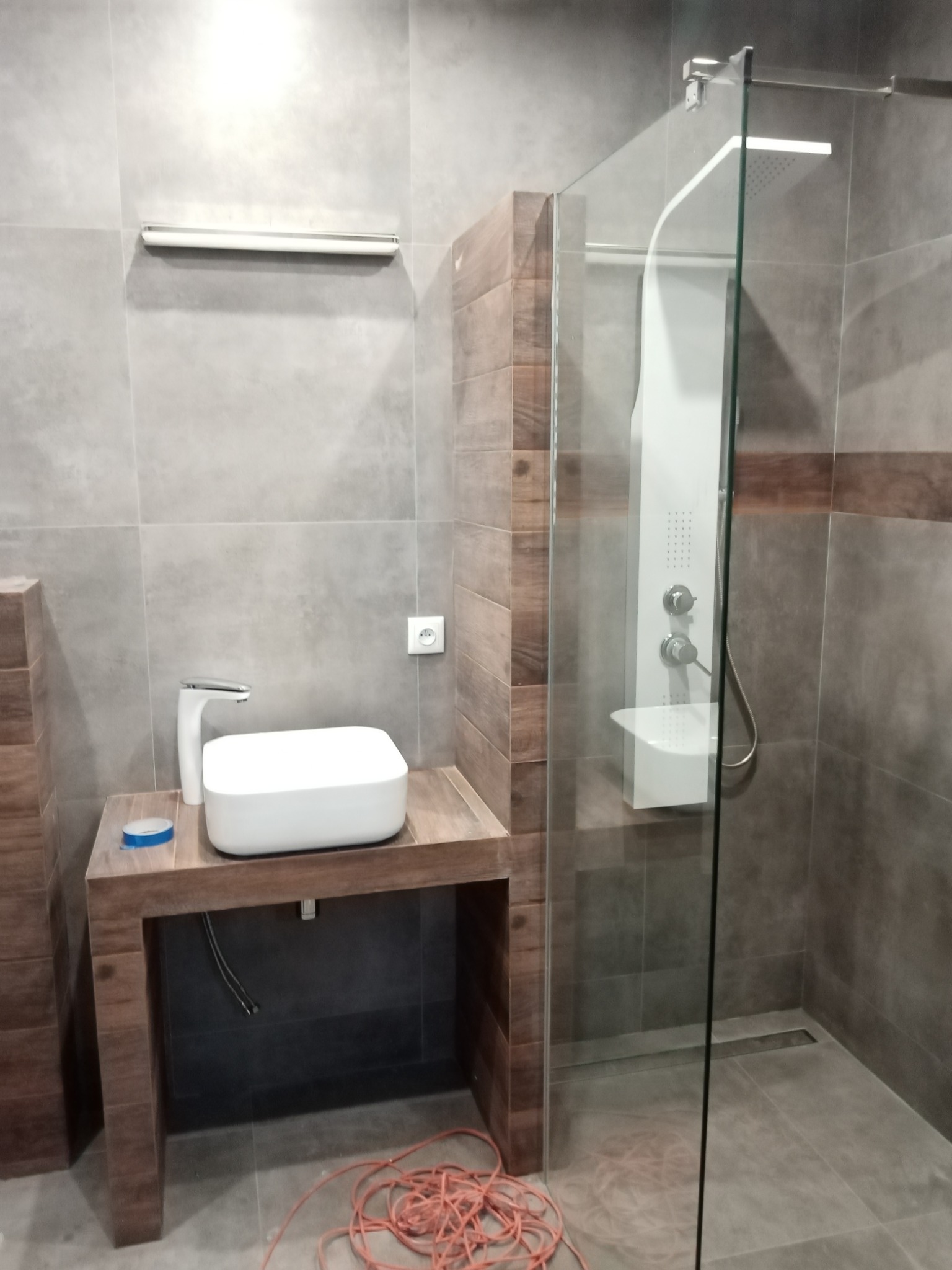 Nowoczesna łazienka z szarą glazurą, drewnopodobnymi akcentami, kabiną prysznicową typu walk-in z panelem natryskowym, białą umywalką nablatową na drewnianej konsoli i widocznym kablem elektrycznym...