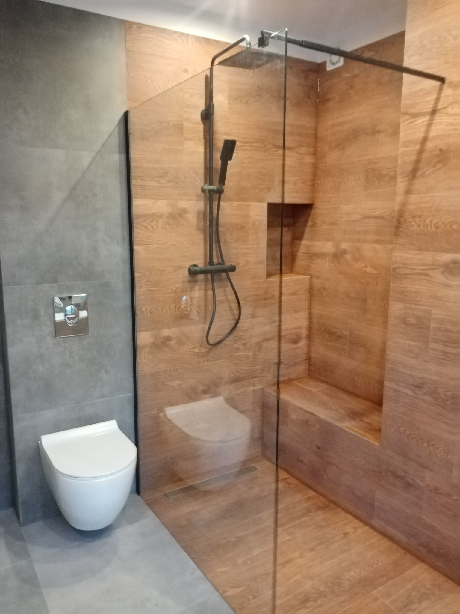 Nowoczesna łazienka z kabiną prysznicową typu walk-in, wykończona płytkami imitującymi drewno i szarymi płytkami, z podtynkową armaturą i toaletą bezkołnierzową.