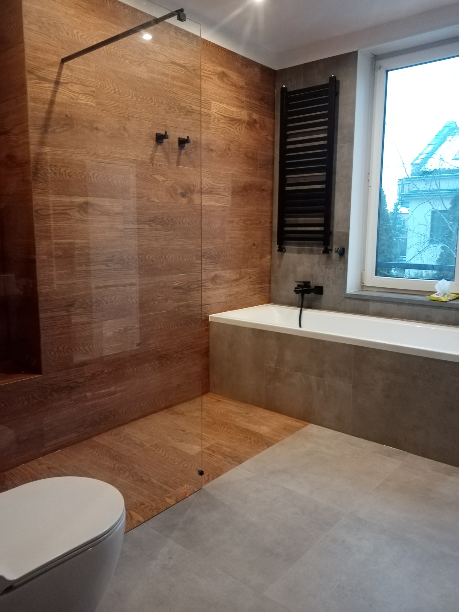 Nowoczesna łazienka z drewnopodobnymi płytkami na ścianach prysznica typu walk-in, szarymi płytkami podłogowymi, białą wanną obudowaną szarymi płytkami oraz czarnym grzejnikiem drabinkowym