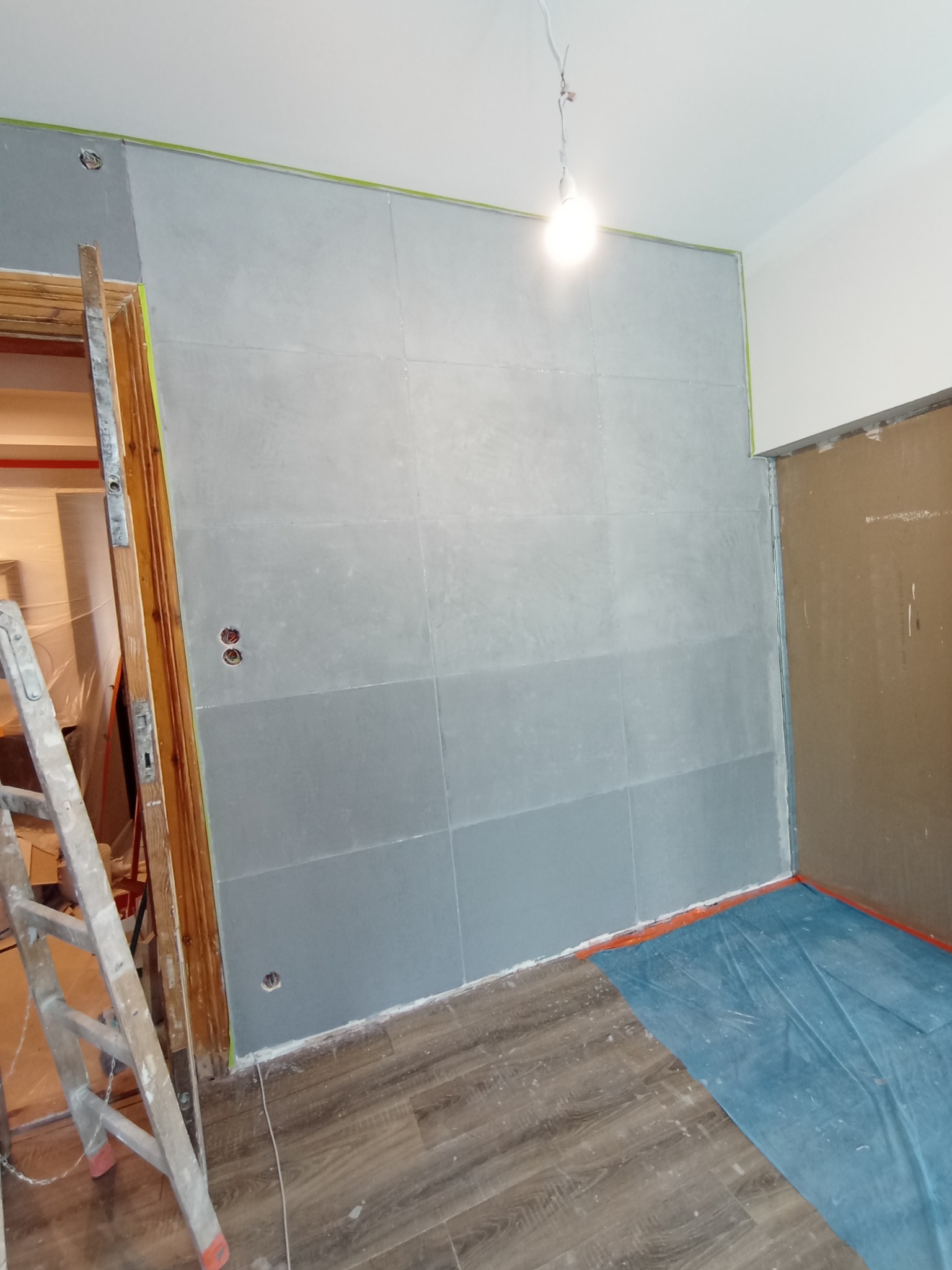 Szare płytki imitujące beton ułożone na ścianie w trakcie remontu, widoczne zabezpieczenia folią i drabina malarska.