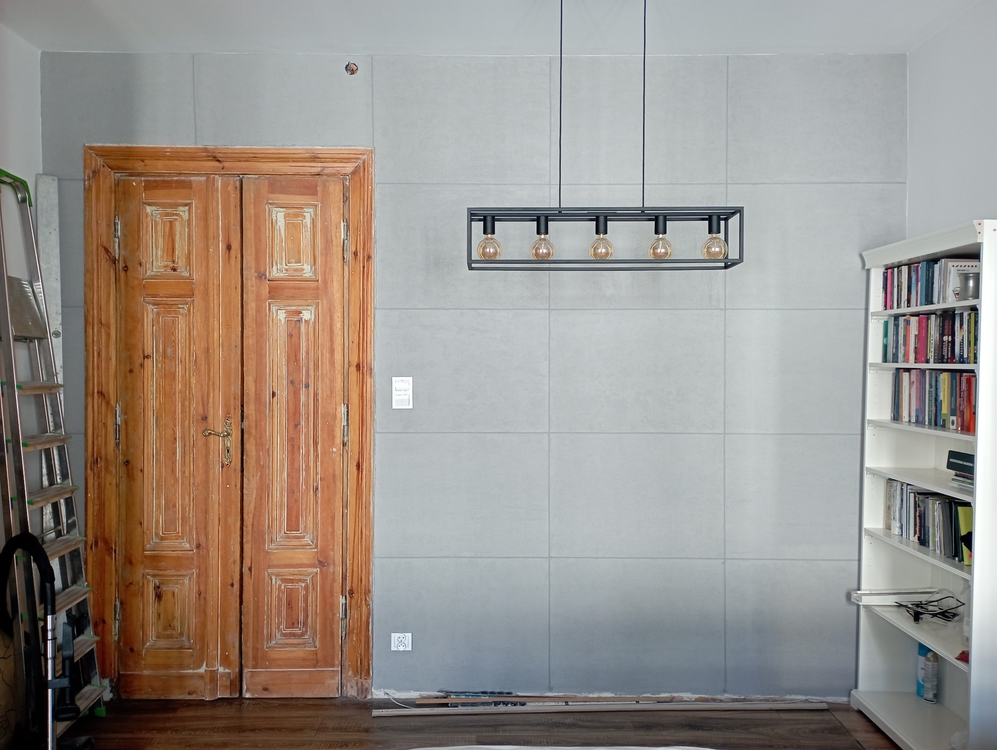 Wnętrze w trakcie prac w Warszawie: drewniane, dwuskrzydłowe drzwi z dekoracyjnymi płycinami, szara ściana z widocznymi gniazdkami, czarna, podwieszana lampa z żarówkami Edisona, biały regał...