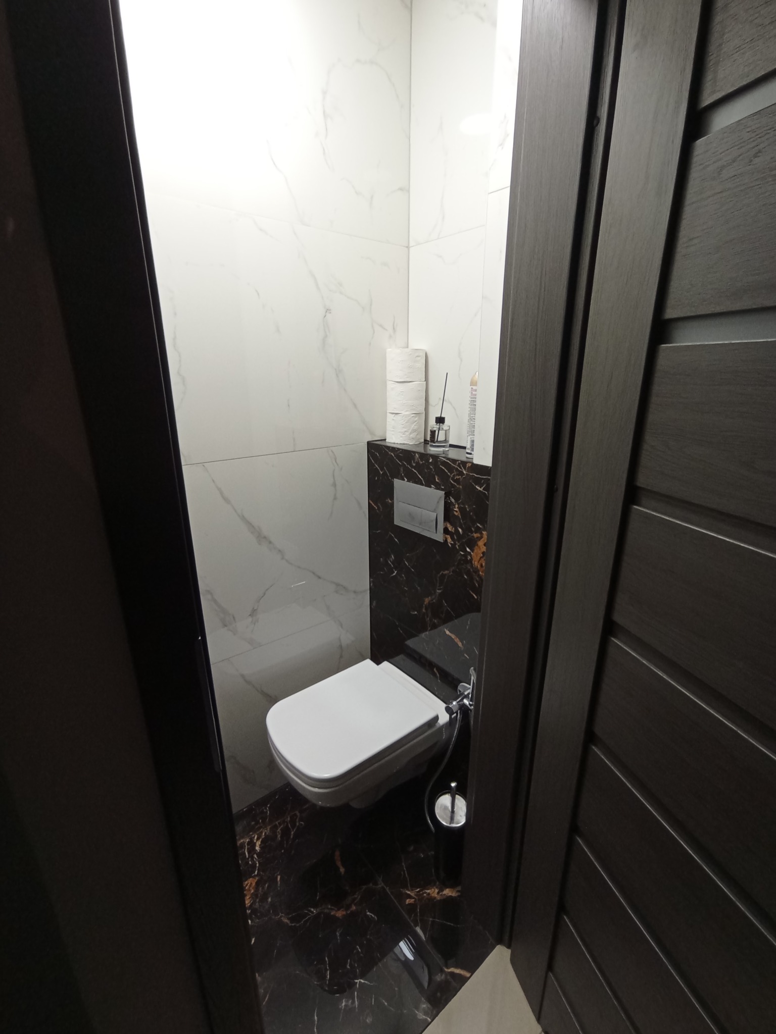 Nowoczesna toaleta podwieszana z bidetką, marmurowe płytki na ścianach i podłodze, widok zza ciemnych drewnianych drzwi.