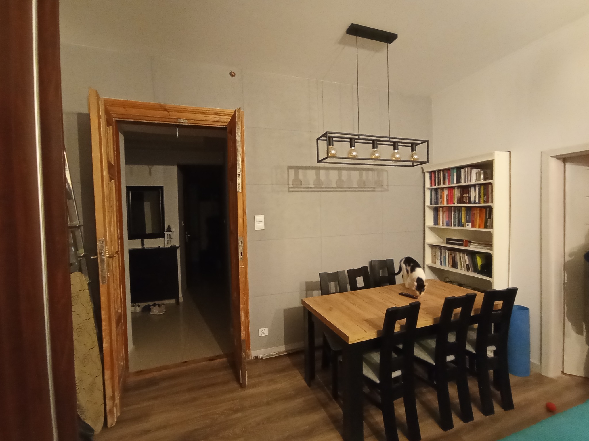 Pokój z drewnianym stołem i czarnymi krzesłami, kot na stole, otwarta drewniana rama drzwiowa, szare płytki na ścianie, wisząca lampa z żarówkami w stylu Edison