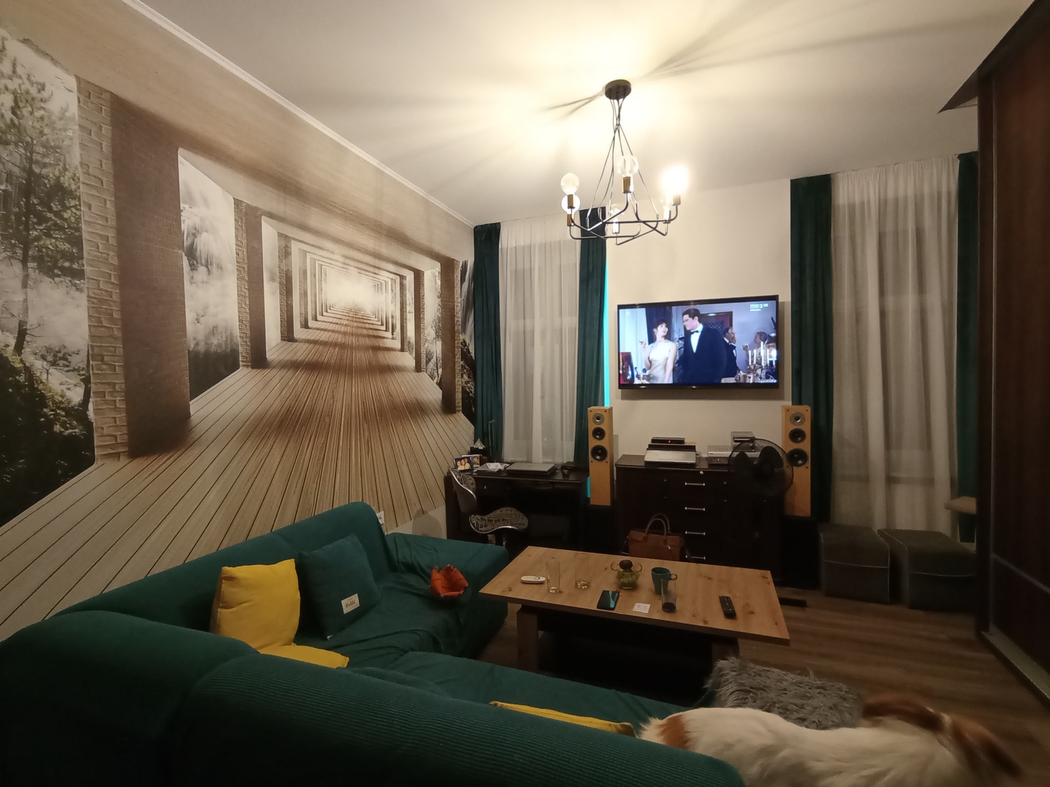 Salon z zieloną sofą, fototapetą z motywem tunelu, drewnianym stolikiem kawowym i wiszącą lampą; na ścianie telewizor z wyświetlanym filmem.