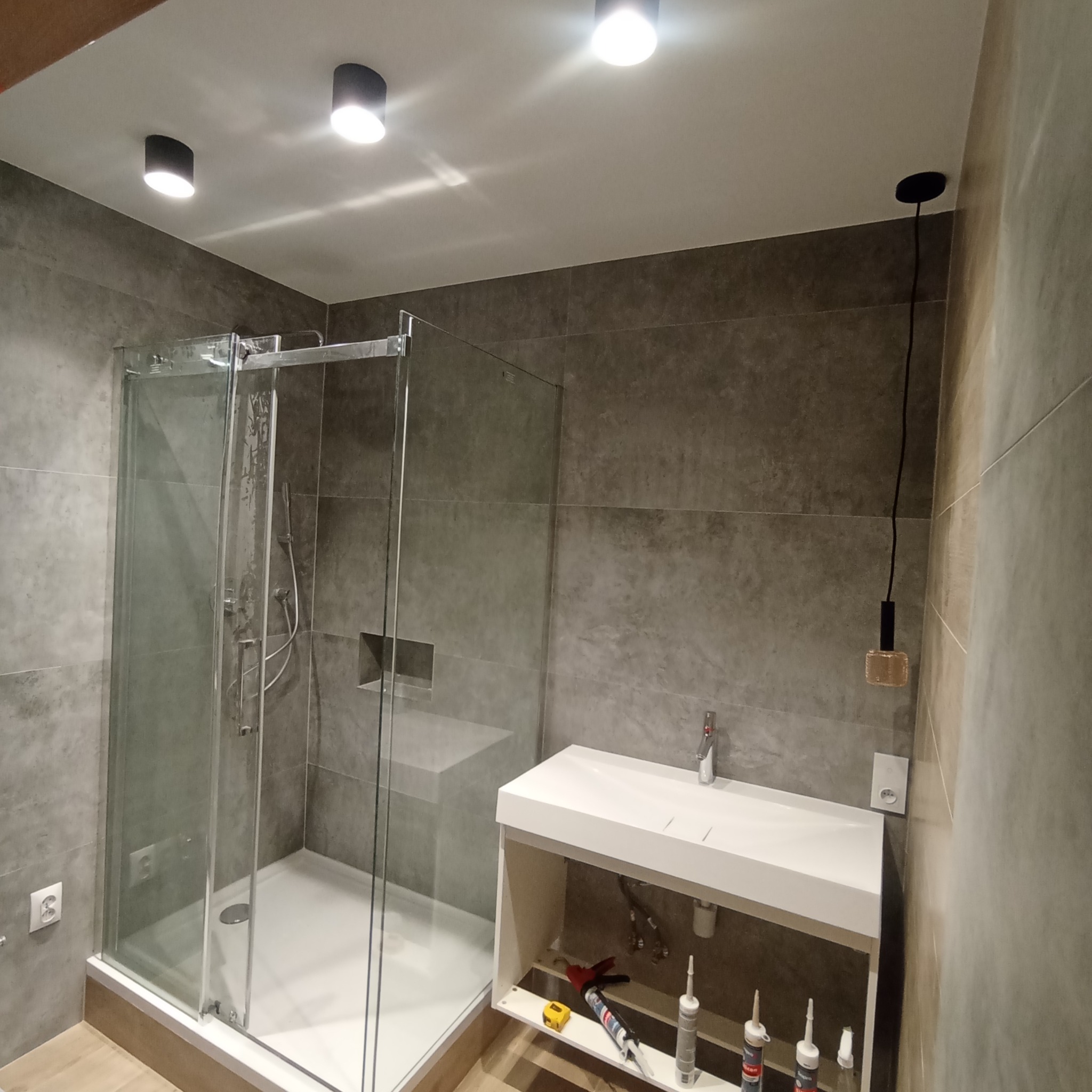 Wykończona łazienka z kabiną prysznicową, umywalką z szafką i szarą glazurą na ścianach, widoczne narzędzia i silikony pod umywalką.