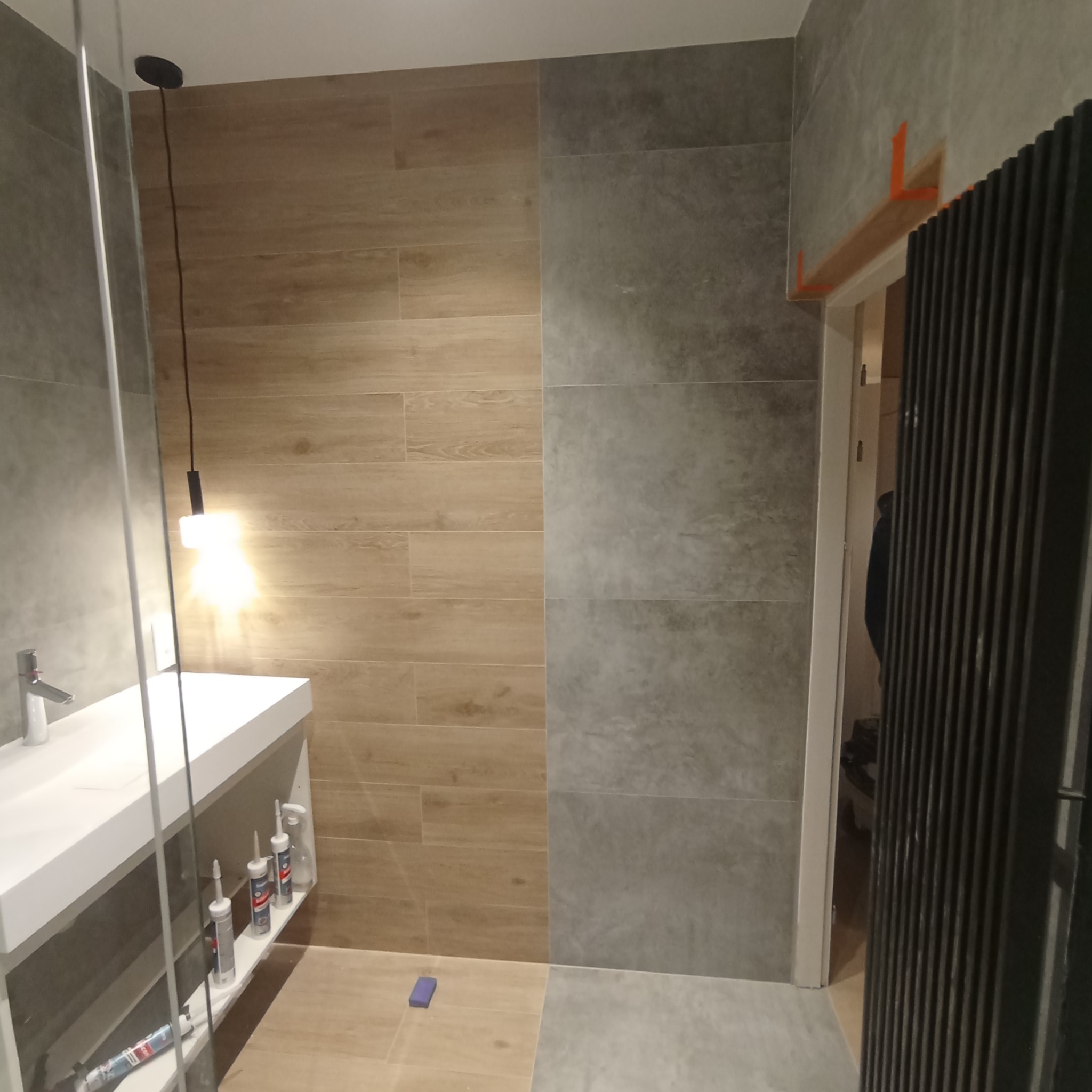 Nowoczesna łazienka w trakcie wykańczania: drewnopodobne i szare płytki, biała umywalka z silikonami na półce, czarny grzejnik, widoczne listwy montażowe nad drzwiami.