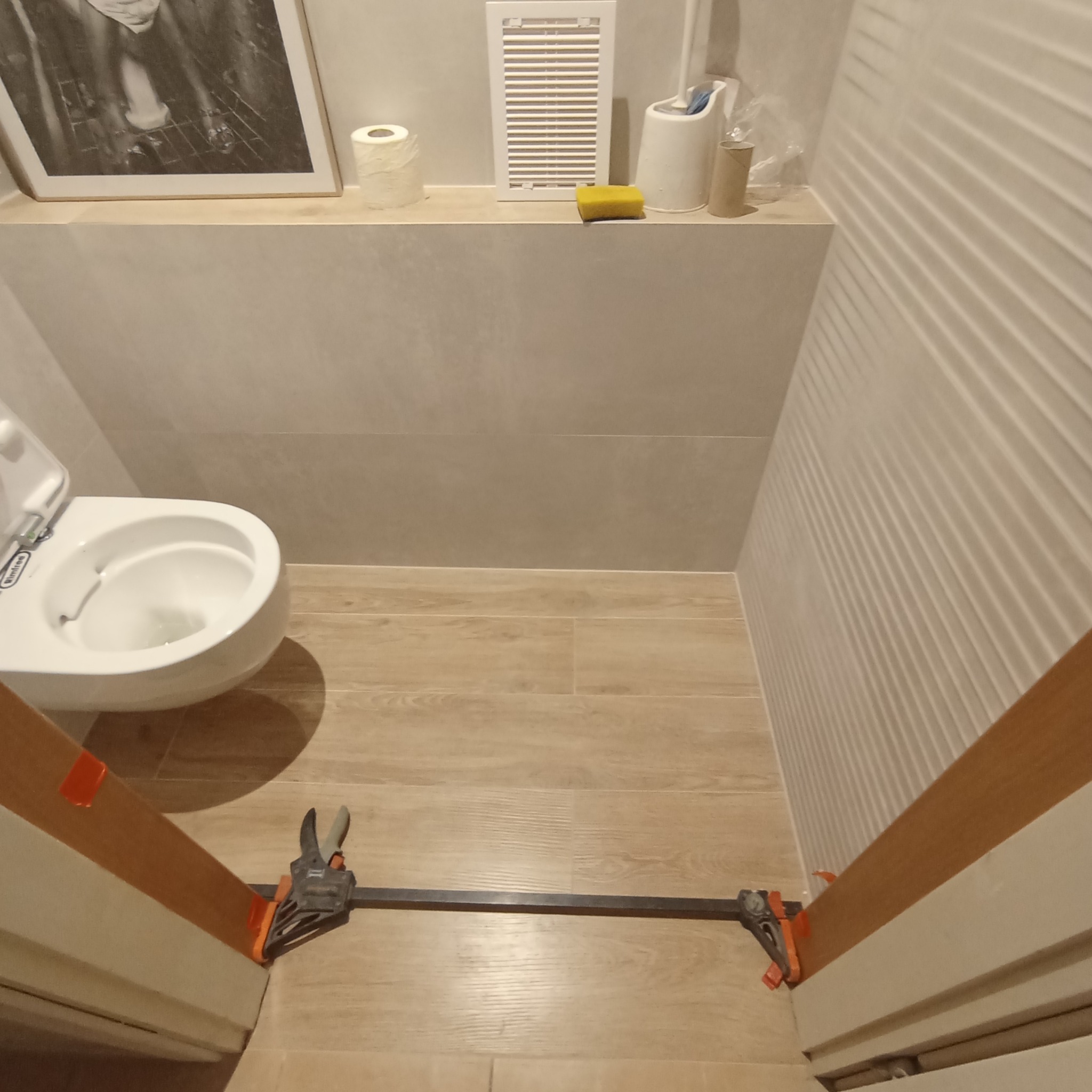 Biała toaleta z otwartą klapą w trakcie prac wykończeniowych, widoczne płytki imitujące drewno na podłodze i dekoracyjne płytki na ścianie, zabezpieczone klamrami stolarskimi.