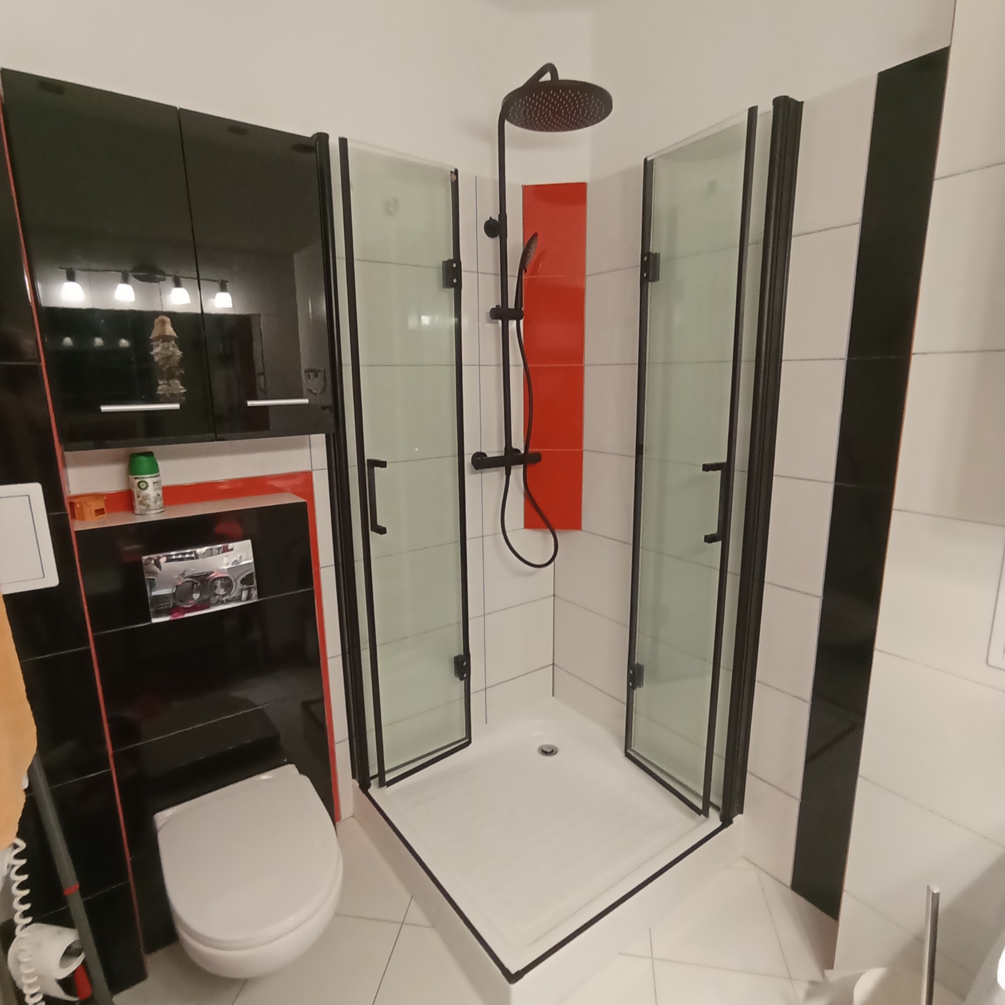 Nowoczesna kabina prysznicowa z czarnymi ramami i deszczownicą w wyremontowanej łazience z czarno-białymi płytkami i czerwonym akcentem na ścianie.