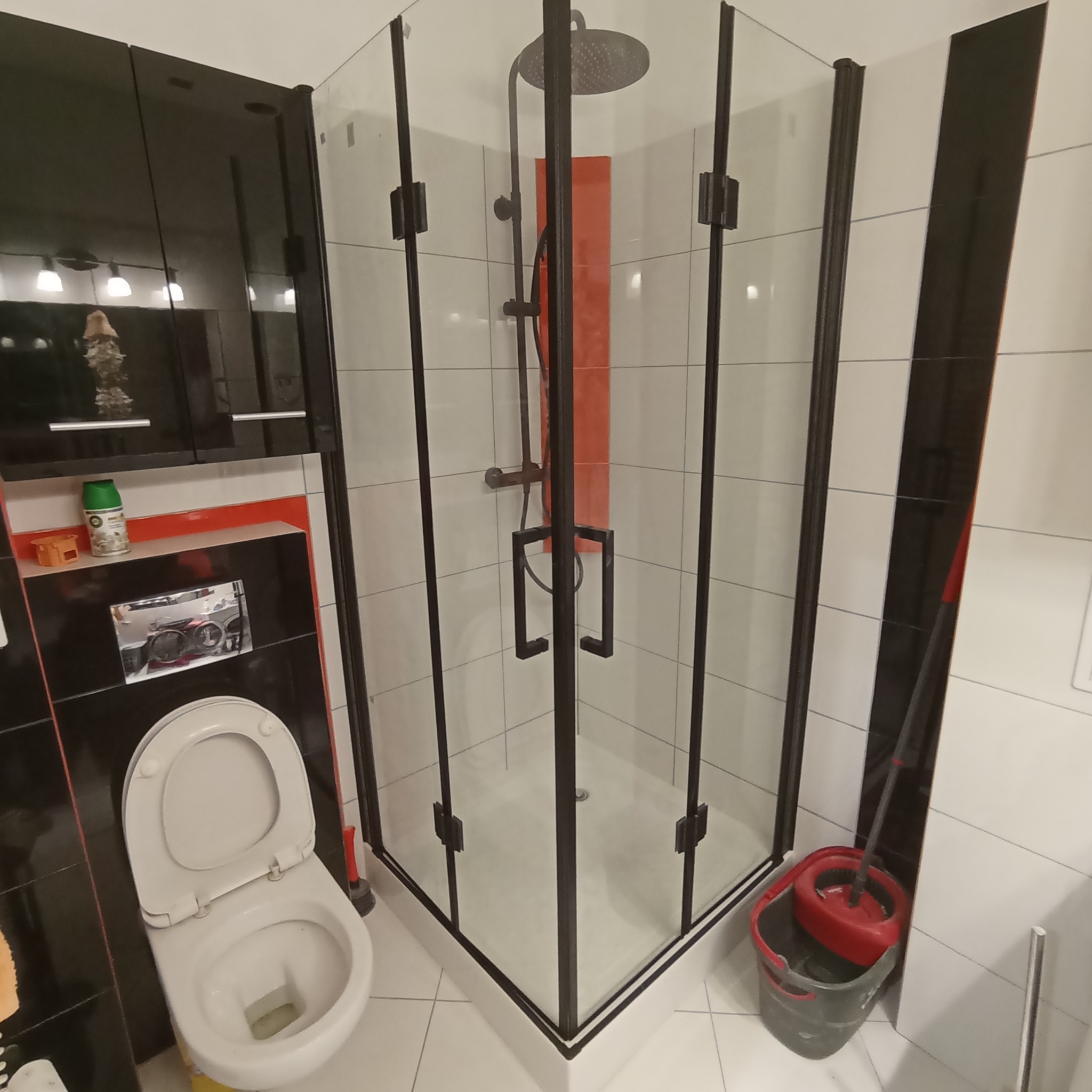 Wykończona łazienka z kabiną prysznicową z czarnymi ramami, wisząca toaleta z otwartą klapą, mop z wiadrem w rogu, płytki w odcieniach bieli i czerni, akcesoria łazienkowe na półkach.