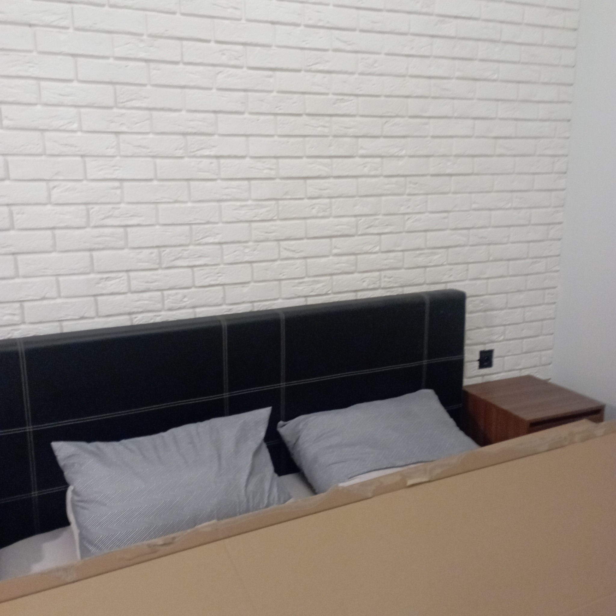 Ściana wykończona dekoracyjną białą cegłą, widoczny fragment łóżka z czarnym zagłówkiem i szarymi poduszkami oraz drewniana szafka nocna
