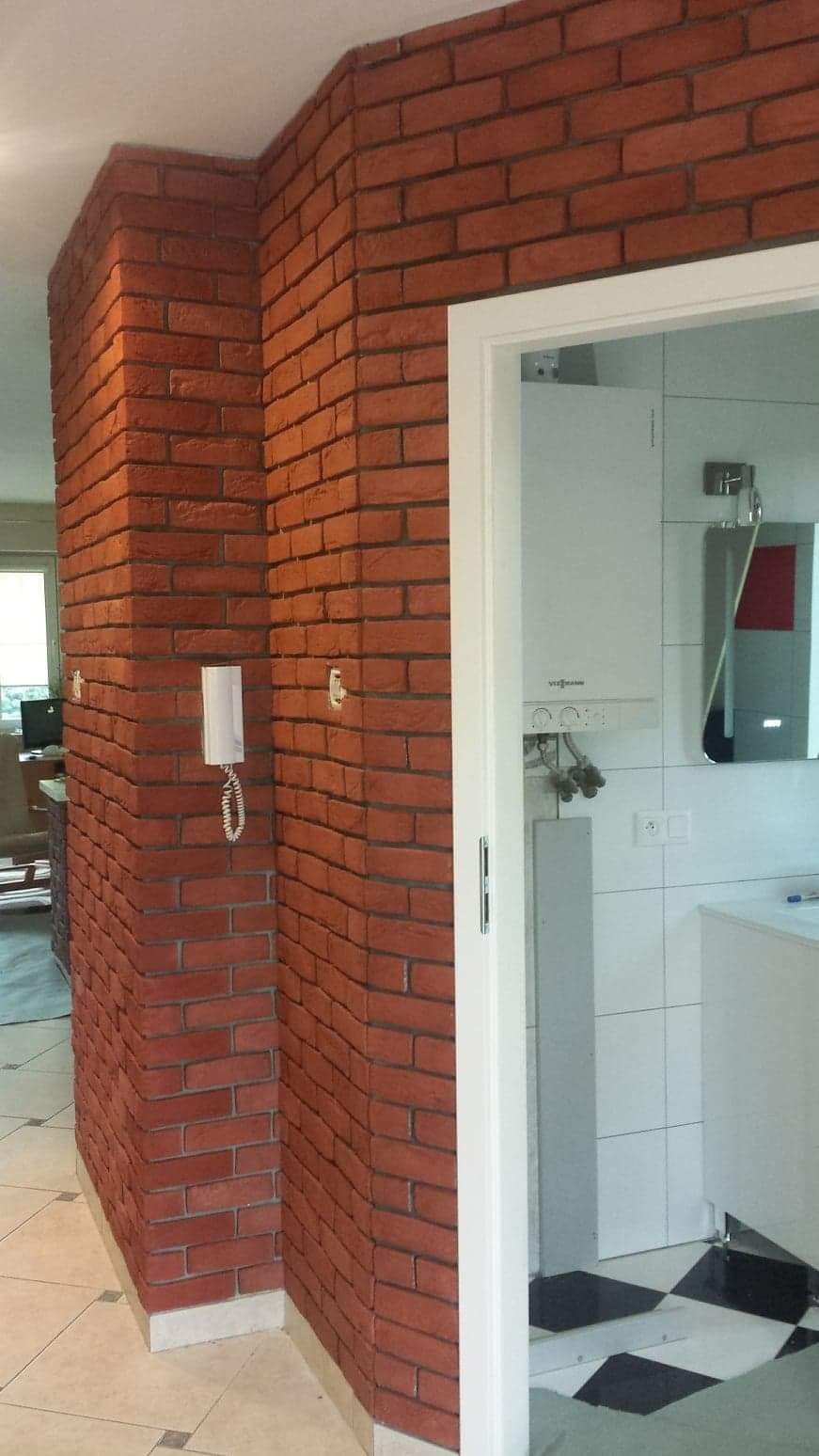 Ściana wykończona czerwoną cegłą klinkierową, z widocznym narożnikiem i fragmentem łazienki z piecem gazowym Viessmann w tle.