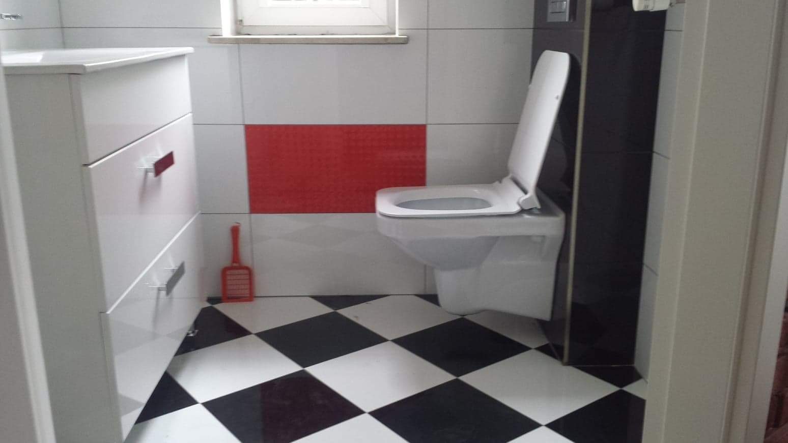 Nowoczesna łazienka z białą szafką z szufladami, toaletą z otwartą klapą, czarno-białą podłogą w szachownicę i czerwonym akcentem na ścianie z płytek, widoczna szczotka do WC.