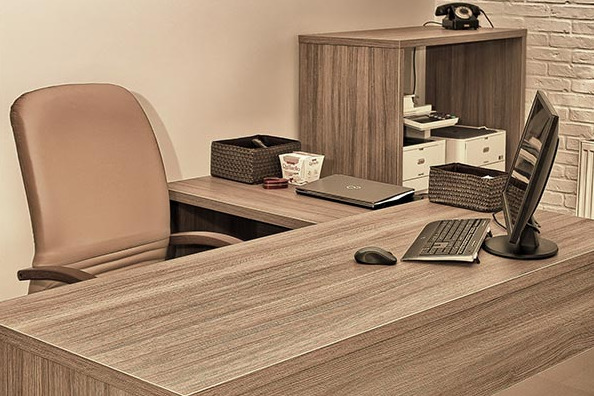 Biuro z drewnianym blatem, komputerem, klawiaturą, myszką, telefonem stacjonarnym na szafce i krzesłem biurowym, utrzymane w ciepłej, stonowanej kolorystyce.