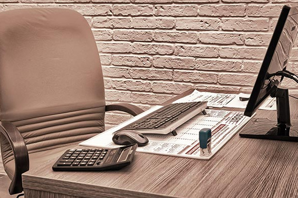 Biurko z kalkulatorem, klawiaturą, myszką, monitorem i stosem dokumentów, pieczątka w tle ściana z cegły