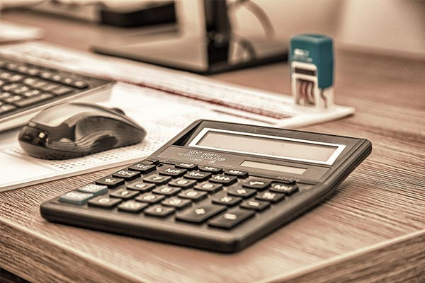 Kalkulator biurowy, myszka i stempel na drewnianym biurku, w tle monitor komputerowy.