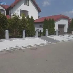 Nowoczesny podjazd i chodnik z jasnoszarej kostki betonowej, obramowane ciemnoszarym pasem, prowadzące do garażu w białym domu z czerwonym dachem. Widoczny słupek ogrodzeniowy i fragment ulicy...