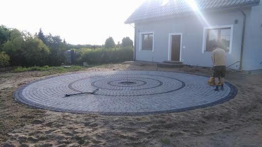Okrągły plac z kostki brukowej ułożony w spiralny wzór przed domem w Świdwinie, pracownik zagęszcza powierzchnię ubijarką.