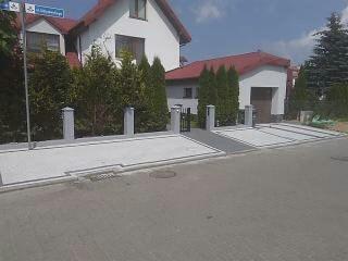 Nowoczesny podjazd i chodnik z jasnoszarej kostki betonowej, obramowane ciemnoszarym pasem, prowadzące do garażu w białym domu z czerwonym dachem. Widoczny słupek ogrodzeniowy i fragment ulicy...