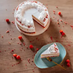 Tort z kremem i malinami na drewnianym stole, z wyciętym kawałkiem i porcją na turkusowym talerzyku z złotym widelcem, posypany drobnymi kawałkami malin.