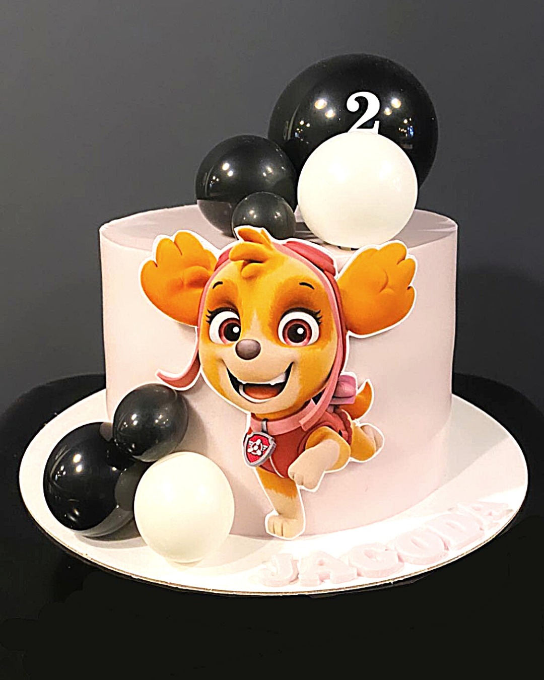 Tort urodzinowy w pastelowych kolorach z figurką Skye z Psiego Patrolu, czarnymi i białymi balonami oraz imieniem 'Jacobo' na podstawie.