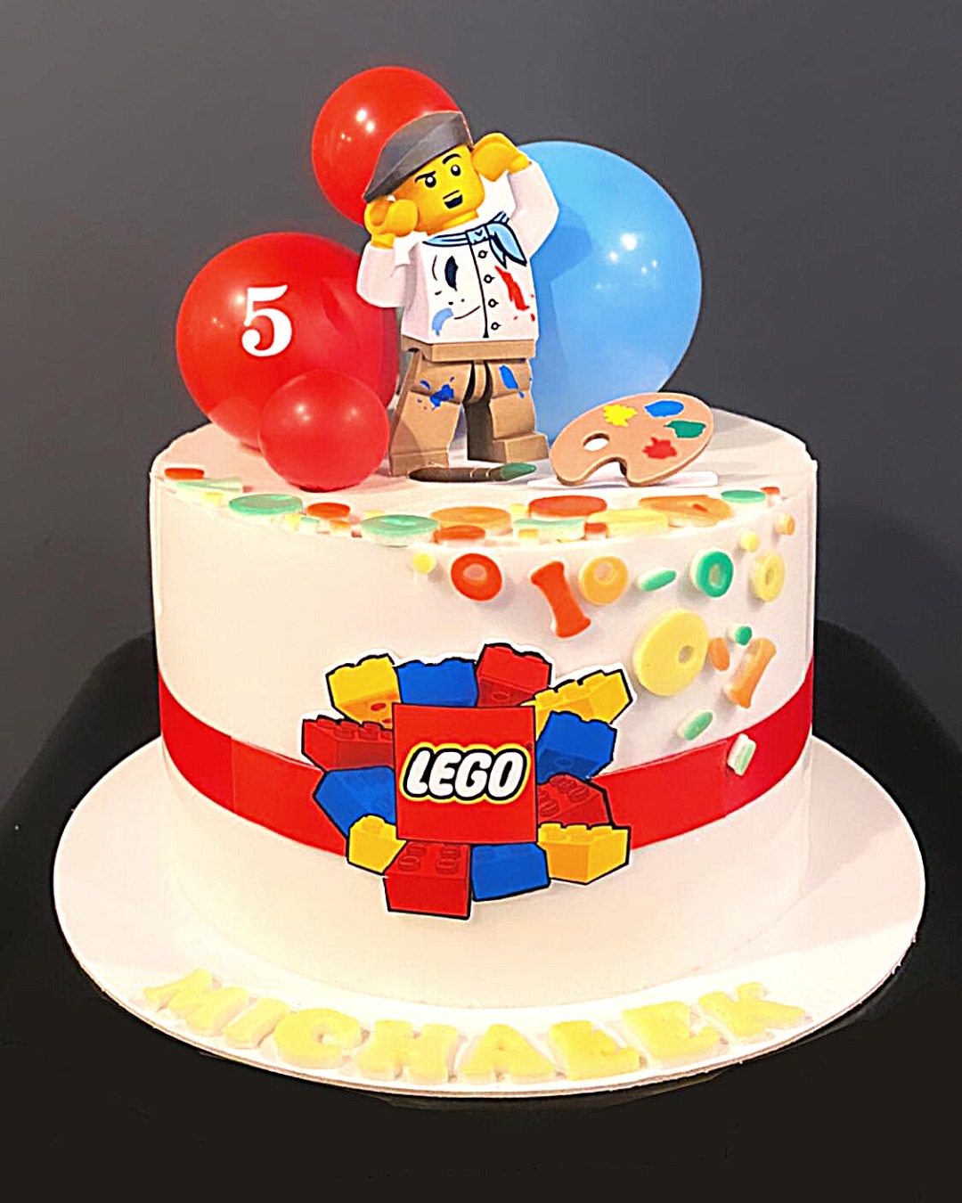 Tort urodzinowy z motywem LEGO: figurka malarza LEGO, balony (jeden z cyfrą '5'), paleta malarska, dekoracje z klocków LEGO i imię 'Michael' z żółtych literek.