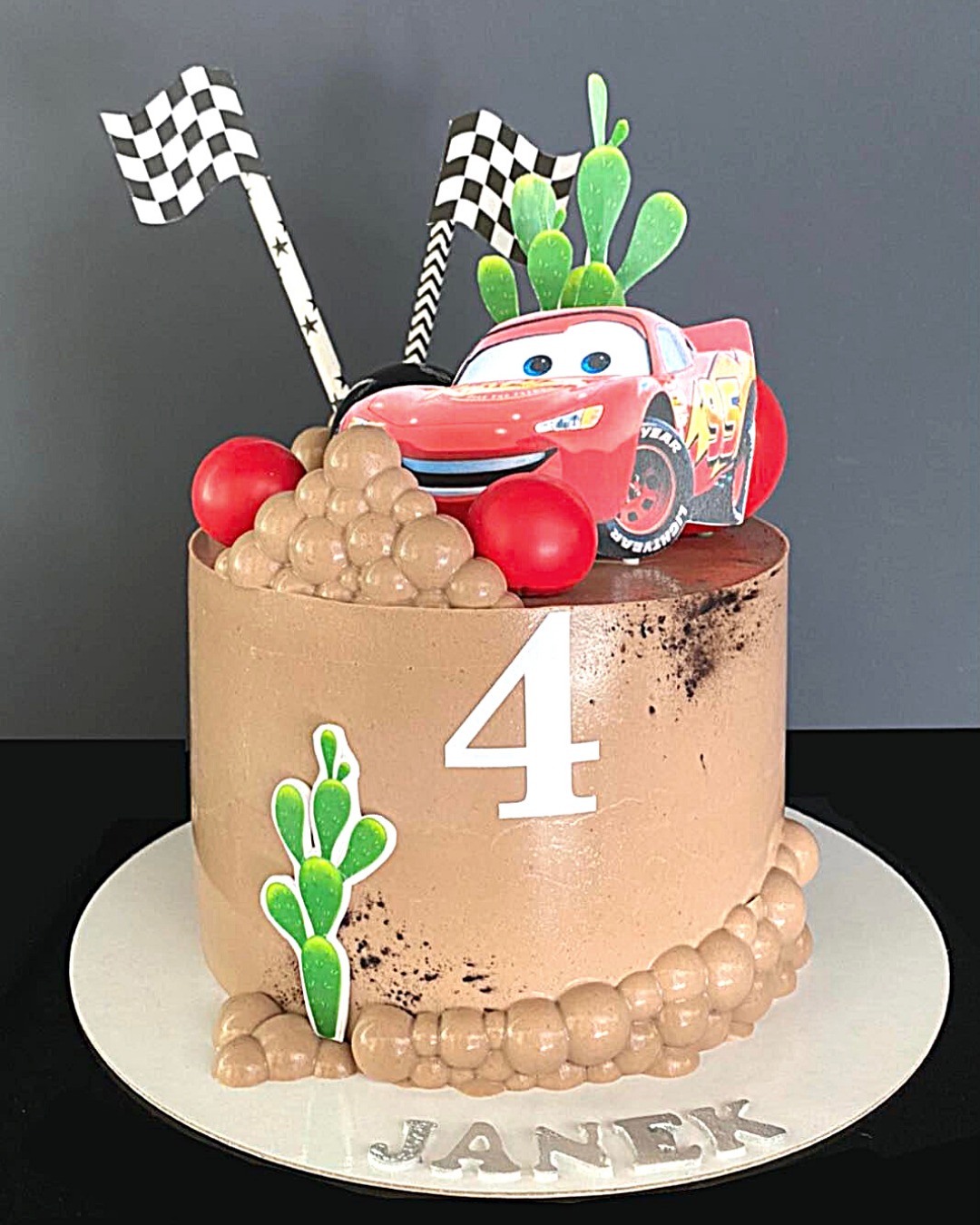 Tort urodzinowy z motywem bajki Auta, dekorowany figurką Zygzaka McQueena, flagami wyścigowymi, kaktusami i liczbą 4, z imieniem Janek na podstawie.