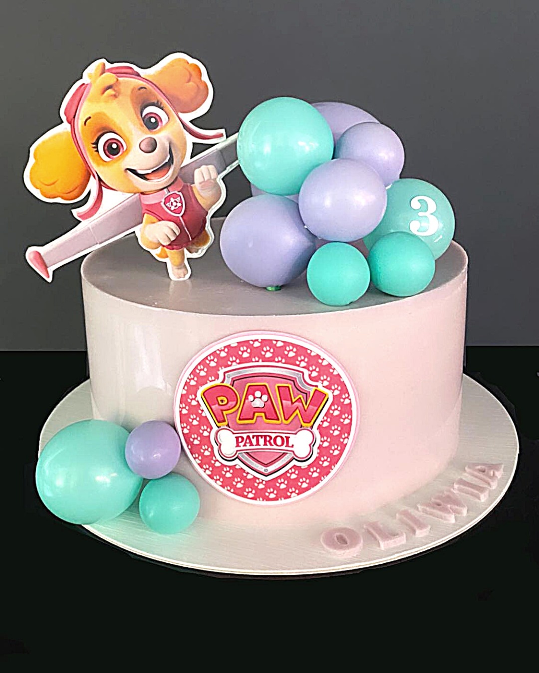 Tort urodzinowy w kolorze różowym, ozdobiony figurką Skye z Psiego Patrolu, balonami w odcieniach fioletu i turkusu, oraz napisem 'Oliwia' z masy cukrowej.